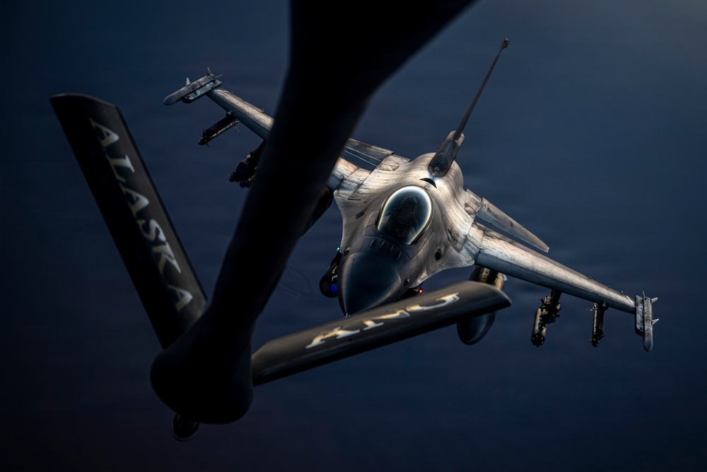 An aerial refueling operation is captured from the perspective of a tanker aircraft, showing a fighter jet approaching the refueling boom. The foreground displays the tanker's tail stabilizer with the word 'ALASKA' inscribed, while an F-16 Fighting Falcon closes in for fuel transfer. The image is dramatically lit with strong contrast, highlighting the fighter's airframe and weapons loadout against a dark background.