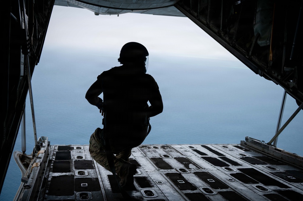 A silhouetted military personnel is crouching at the open rear ramp of a military cargo aircraft in flight, looking out over a large body of water below. The figure is wearing tactical gear including a helmet, body armor, and combat equipment. The image is dramatically backlit, creating a strong silhouette contrast against the blue-grey sky and ocean backdrop.