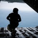 A silhouetted military personnel is crouching at the open rear ramp of a military cargo aircraft in flight, looking out over a large body of water below. The figure is wearing tactical gear including a helmet, body armor, and combat equipment. The image is dramatically backlit, creating a strong silhouette contrast against the blue-grey sky and ocean backdrop.