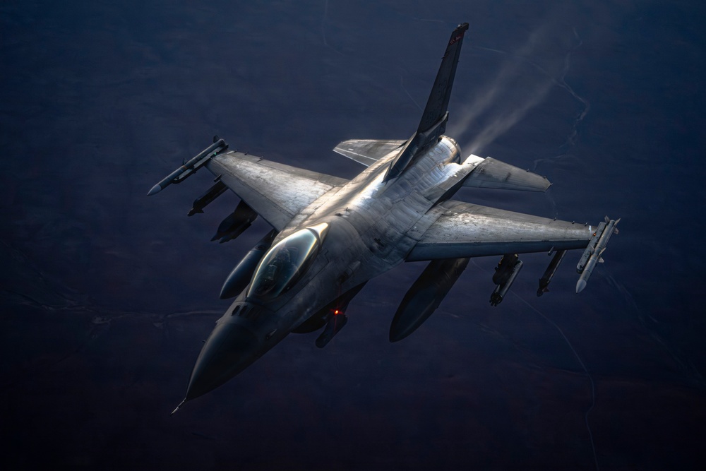 An aerial photograph taken from above showing a single-engine fighter jet in flight over a dark, expansive landscape. The aircraft is illuminated by directional light, highlighting its metallic surface and underwing ordnance. Engine exhaust vapor is visible trailing from the rear of the aircraft.