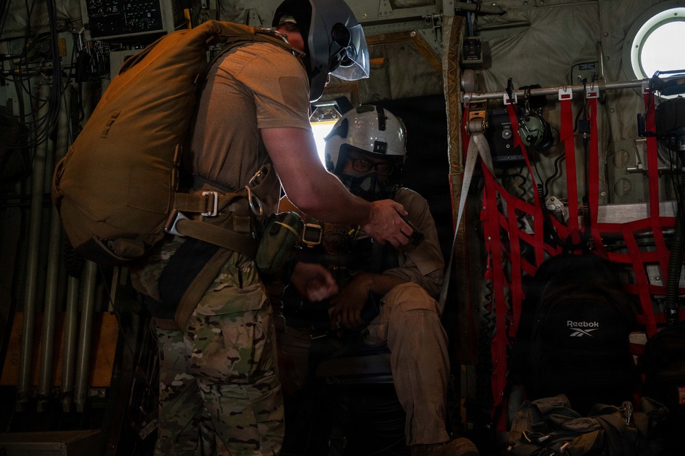 Two military personnel are inside what appears to be a military transport aircraft, likely a C-130 or similar aircraft. One standing individual wearing multicam fatigues and a parachute pack appears to be assisting or checking equipment on a seated individual wearing a flight helmet with visor. The interior shows typical military aircraft features including red cargo netting, wiring, and equipment panels.