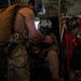 Two military personnel are inside what appears to be a military transport aircraft, likely a C-130 or similar aircraft. One standing individual wearing multicam fatigues and a parachute pack appears to be assisting or checking equipment on a seated individual wearing a flight helmet with visor. The interior shows typical military aircraft features including red cargo netting, wiring, and equipment panels.