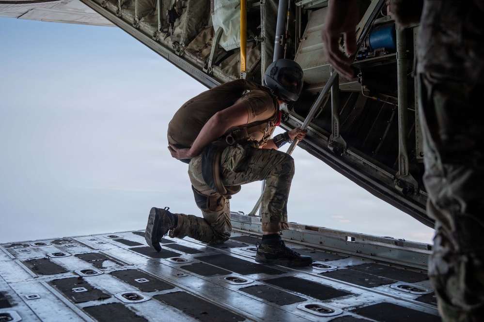 A military personnel wearing camouflage uniform and a flight helmet is crouched on the open rear ramp of a military cargo aircraft in flight. The individual appears to be inspecting or adjusting equipment near the ramp edge, with open sky visible below and behind. A second person in military attire is partially visible on the right side of the frame.