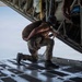 A military personnel wearing camouflage uniform and a flight helmet is crouched on the open rear ramp of a military cargo aircraft in flight. The individual appears to be inspecting or adjusting equipment near the ramp edge, with open sky visible below and behind. A second person in military attire is partially visible on the right side of the frame.