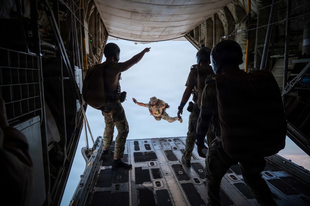 Military personnel are conducting a parachute jump operation from the open rear ramp of a large military cargo aircraft, likely a C-130 Hercules or similar transport. One jumper is already airborne and visible outside the aircraft in freefall position, while several other personnel in full tactical gear stand on the ramp observing and directing. One individual on the left is pointing upward, likely giving a signal or direction.