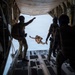 Military personnel are conducting a parachute jump operation from the open rear ramp of a large military cargo aircraft, likely a C-130 Hercules or similar transport. One jumper is already airborne and visible outside the aircraft in freefall position, while several other personnel in full tactical gear stand on the ramp observing and directing. One individual on the left is pointing upward, likely giving a signal or direction.