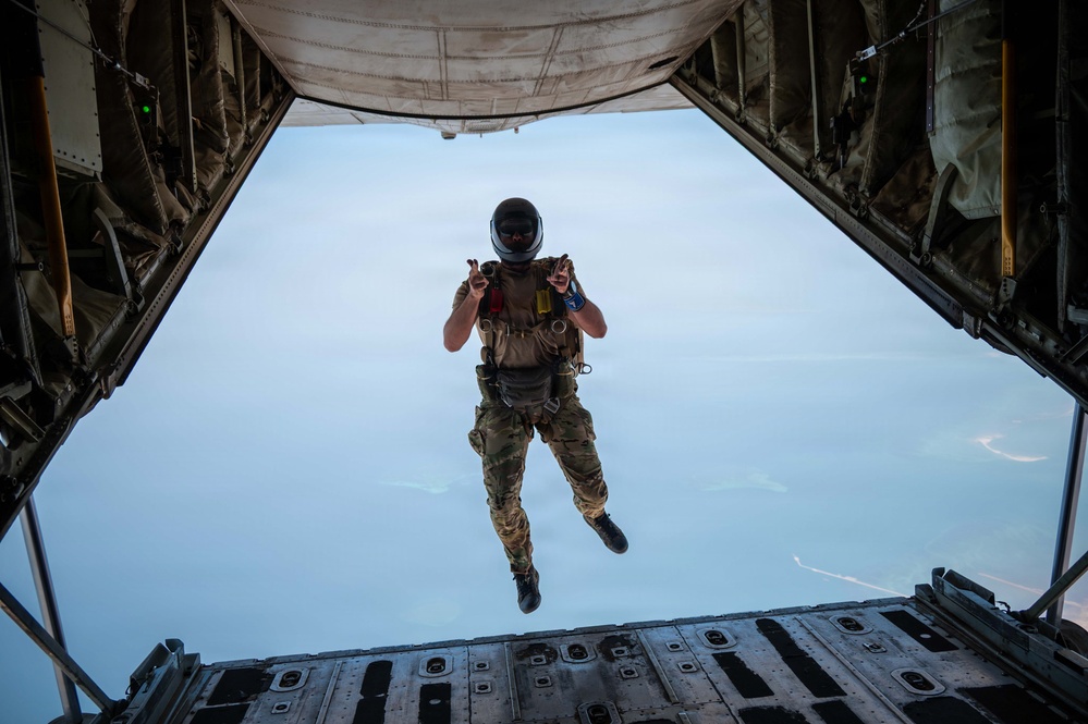 A military parachutist is captured mid-jump exiting the open rear ramp of a large military cargo aircraft, likely a C-130 or C-17. The jumper is wearing full tactical gear including a helmet with visor, multicam uniform, parachute harness, and tactical vest, and appears to be giving a thumbs-up or peace sign gesture. The sky below is clear and blue, with what appears to be a coastline or water visible in the distant background.