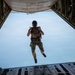 A military parachutist is captured mid-jump exiting the open rear ramp of a large military cargo aircraft, likely a C-130 or C-17. The jumper is wearing full tactical gear including a helmet with visor, multicam uniform, parachute harness, and tactical vest, and appears to be giving a thumbs-up or peace sign gesture. The sky below is clear and blue, with what appears to be a coastline or water visible in the distant background.