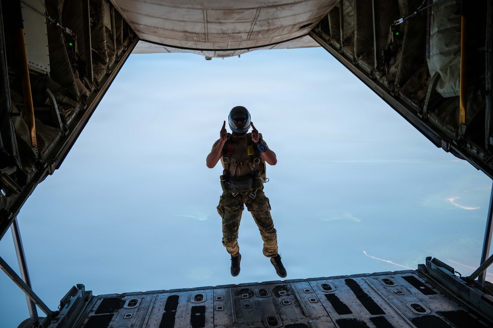 A military paratrooper is captured mid-jump from the open rear ramp of a large military cargo aircraft, likely a C-130 Hercules or similar transport. The jumper is wearing full tactical gear including a helmet with visor, multicam fatigues, parachute harness, and tactical vest, and appears to be giving a hand signal. The sky below is hazy and blue, with faint landmasses or contrails visible in the background.