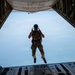 A military paratrooper is captured mid-jump from the open rear ramp of a large military cargo aircraft, likely a C-130 Hercules or similar transport. The jumper is wearing full tactical gear including a helmet with visor, multicam fatigues, parachute harness, and tactical vest, and appears to be giving a hand signal. The sky below is hazy and blue, with faint landmasses or contrails visible in the background.