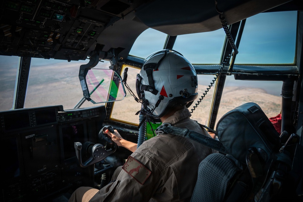 A military pilot is seated in the cockpit of a large aircraft, viewed from behind, actively flying over an arid, semi-desert landscape. The pilot is wearing a flight helmet with red triangle markings and a flight suit with a visible patch on the shoulder. Through the cockpit windows, another aircraft with green and pink coloring is visible in close proximity, suggesting a formation flight or aerial refueling operation.