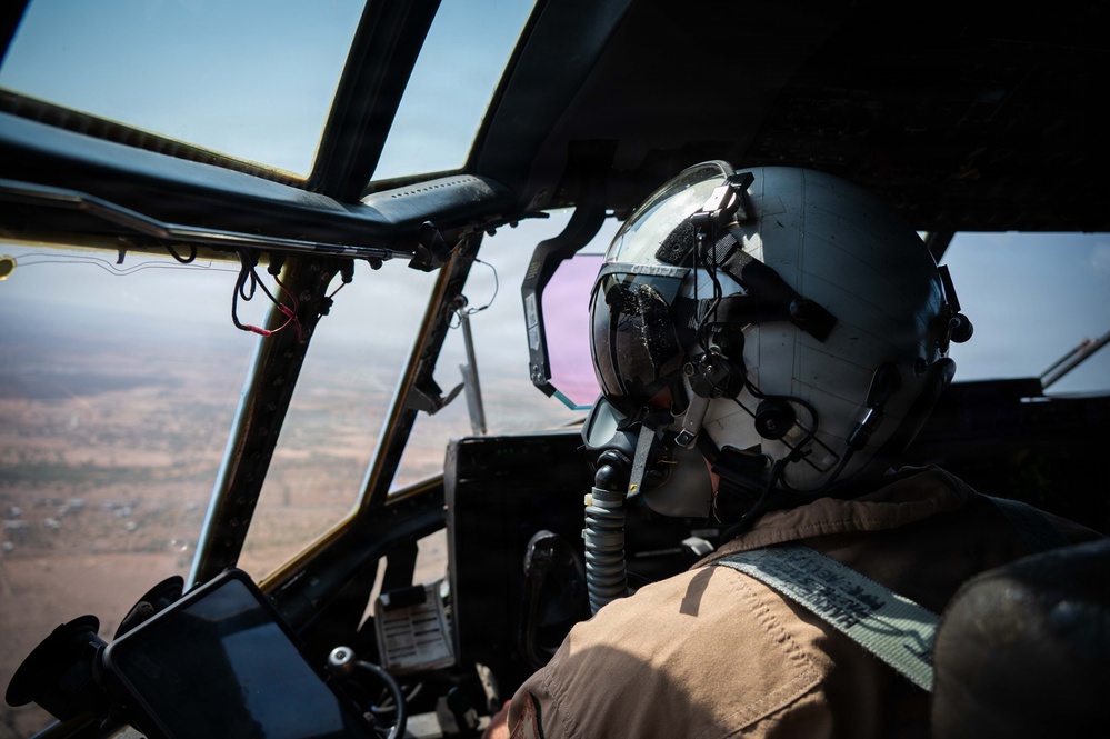 A military pilot is seated in the cockpit of a large aircraft, viewed from behind and slightly to the side. The pilot is wearing a full flight helmet with a tinted visor, oxygen mask, and tan/desert flight suit. The aircraft is airborne over what appears to be an arid, semi-urban landscape visible through the large cockpit windows.