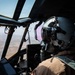 A military pilot is seated in the cockpit of a large aircraft, viewed from behind and slightly to the side. The pilot is wearing a full flight helmet with a tinted visor, oxygen mask, and tan/desert flight suit. The aircraft is airborne over what appears to be an arid, semi-urban landscape visible through the large cockpit windows.