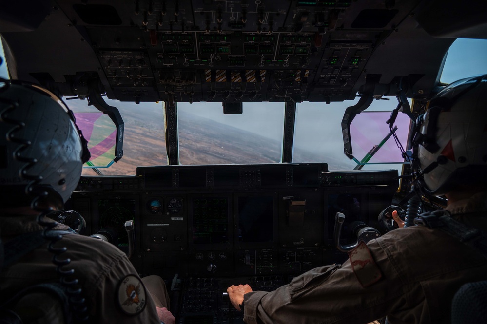 Two military aircrew members are visible from behind in the cockpit of a large military transport aircraft, flying over mountainous or arid terrain. The cockpit features an extensive array of instruments, displays, and overhead panels consistent with a large multi-engine military transport such as a C-130 or C-17. Pink/magenta-tinted side windows and hazy atmospheric conditions are visible through the windscreen.