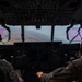 Two military aircrew members are visible from behind in the cockpit of a large military transport aircraft, flying over mountainous or arid terrain. The cockpit features an extensive array of instruments, displays, and overhead panels consistent with a large multi-engine military transport such as a C-130 or C-17. Pink/magenta-tinted side windows and hazy atmospheric conditions are visible through the windscreen.