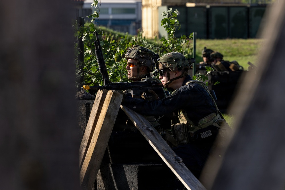 The image shows multiple military personnel in tactical gear taking cover behind a wooden barrier or barricade. At least one soldier is aiming a rifle over the barrier while others observe or provide support. The setting appears to be a training exercise or operational scenario in an urban or semi-urban environment.