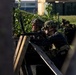 The image shows multiple military personnel in tactical gear taking cover behind a wooden barrier or barricade. At least one soldier is aiming a rifle over the barrier while others observe or provide support. The setting appears to be a training exercise or operational scenario in an urban or semi-urban environment.