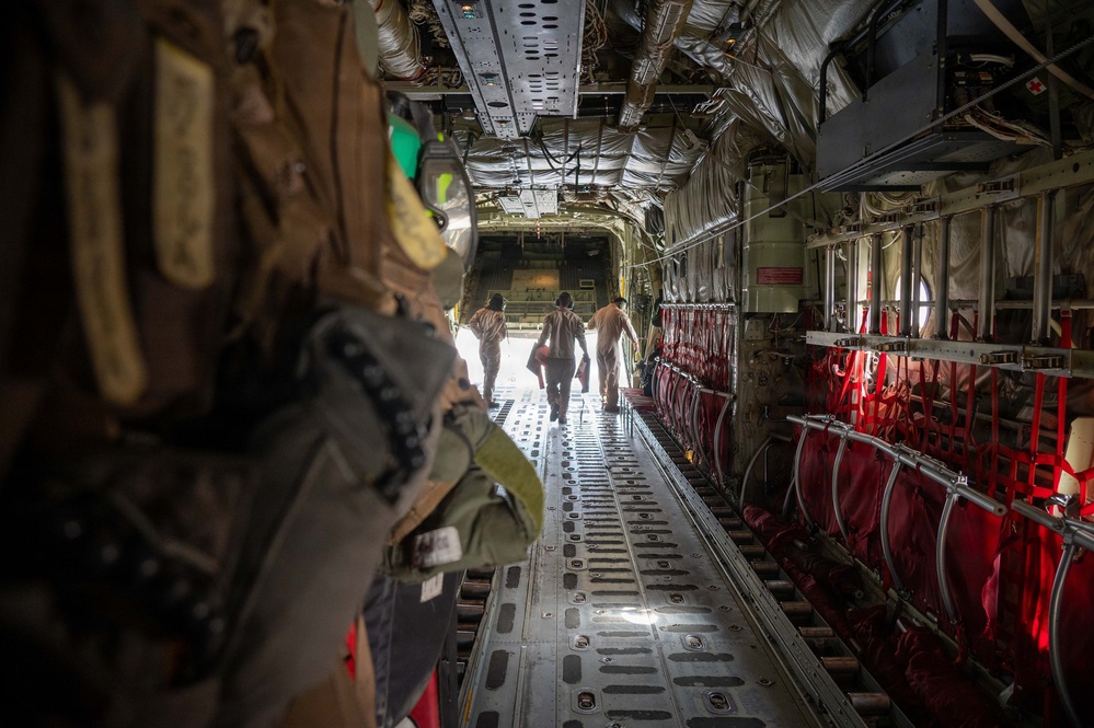 The image shows the interior of a military transport aircraft, likely a C-130 Hercules, viewed from the rear cargo area looking toward the open ramp. Three personnel are walking toward the open rear ramp, silhouetted against bright exterior light. Red cargo netting/seating is visible along the right fuselage wall, and military gear is hanging on the left foreground.