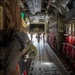 The image shows the interior of a military transport aircraft, likely a C-130 Hercules, viewed from the rear cargo area looking toward the open ramp. Three personnel are walking toward the open rear ramp, silhouetted against bright exterior light. Red cargo netting/seating is visible along the right fuselage wall, and military gear is hanging on the left foreground.