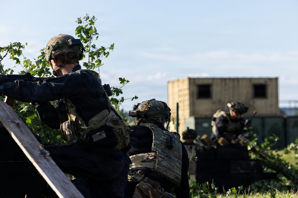 The image depicts military personnel in tactical gear taking covered positions behind a wooden barrier and vegetation. At least three soldiers are visible, with the foreground soldier aiming a rifle while others maintain defensive postures. A military-style modular building or shipping container structure is visible in the background, suggesting a training exercise or operational scenario.