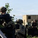 The image depicts military personnel in tactical gear taking covered positions behind a wooden barrier and vegetation. At least three soldiers are visible, with the foreground soldier aiming a rifle while others maintain defensive postures. A military-style modular building or shipping container structure is visible in the background, suggesting a training exercise or operational scenario.