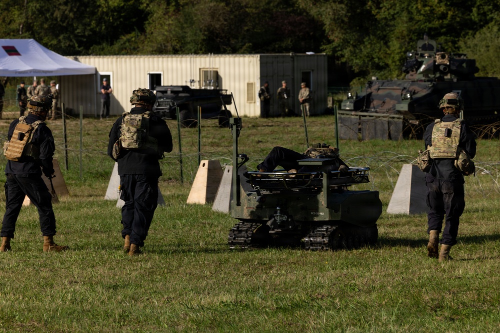 Military personnel in tactical gear are observing or operating a small tracked unmanned ground vehicle (UGV) on a grassy field during what appears to be a training exercise or demonstration. The scene includes concrete barriers, razor wire, armored vehicles in the background, and a command tent area. Multiple soldiers wearing body armor and multicam/black uniforms are present.