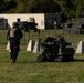 Military personnel in tactical gear are observing or operating a small tracked unmanned ground vehicle (UGV) on a grassy field during what appears to be a training exercise or demonstration. The scene includes concrete barriers, razor wire, armored vehicles in the background, and a command tent area. Multiple soldiers wearing body armor and multicam/black uniforms are present.