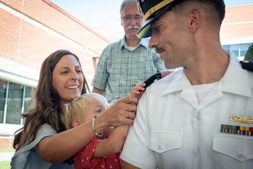 A military officer in a white dress uniform is having insignia or rank devices pinned onto his collar by a smiling woman holding a young blonde child. An older man in a plaid shirt observes the ceremony in the background. This appears to be a military promotion or commissioning ceremony with family members participating.