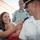 A military officer in a white dress uniform is having insignia or rank devices pinned onto his collar by a smiling woman holding a young blonde child. An older man in a plaid shirt observes the ceremony in the background. This appears to be a military promotion or commissioning ceremony with family members participating.