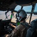 A military pilot is seated in the cockpit of a large aircraft, viewed from behind, actively flying over an arid, semi-desert landscape. The pilot is wearing a flight helmet with red triangle markings and a flight suit with a visible patch on the shoulder. Through the cockpit windows, another aircraft with green and pink coloring is visible in close proximity, suggesting a formation flight or aerial refueling operation.