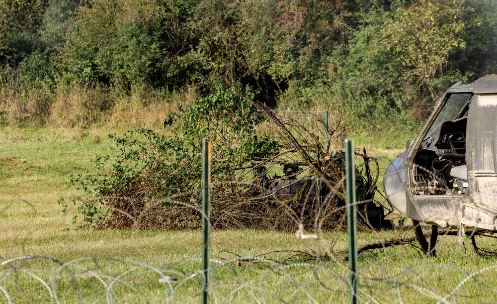 The image shows an abandoned or wrecked helicopter fuselage on a grassy field, partially obscured by fallen branches and overgrown vegetation. A coiled razor wire (concertina wire) fence is visible in the foreground, suggesting a secured or restricted perimeter. The scene appears to be a decommissioned or crash site area with significant vegetation overgrowth.