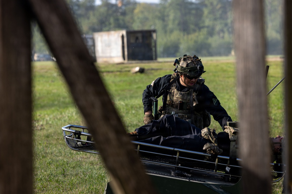 A soldier in full tactical gear and camouflage helmet is attending to a person lying on a military stretcher or litter carrier in an open field setting. The scene appears to depict a combat casualty care or medical evacuation training exercise. The image is captured through a partially obscured foreground, suggesting the photographer is positioned behind a wooden structure.