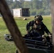 A soldier in full tactical gear and camouflage helmet is attending to a person lying on a military stretcher or litter carrier in an open field setting. The scene appears to depict a combat casualty care or medical evacuation training exercise. The image is captured through a partially obscured foreground, suggesting the photographer is positioned behind a wooden structure.