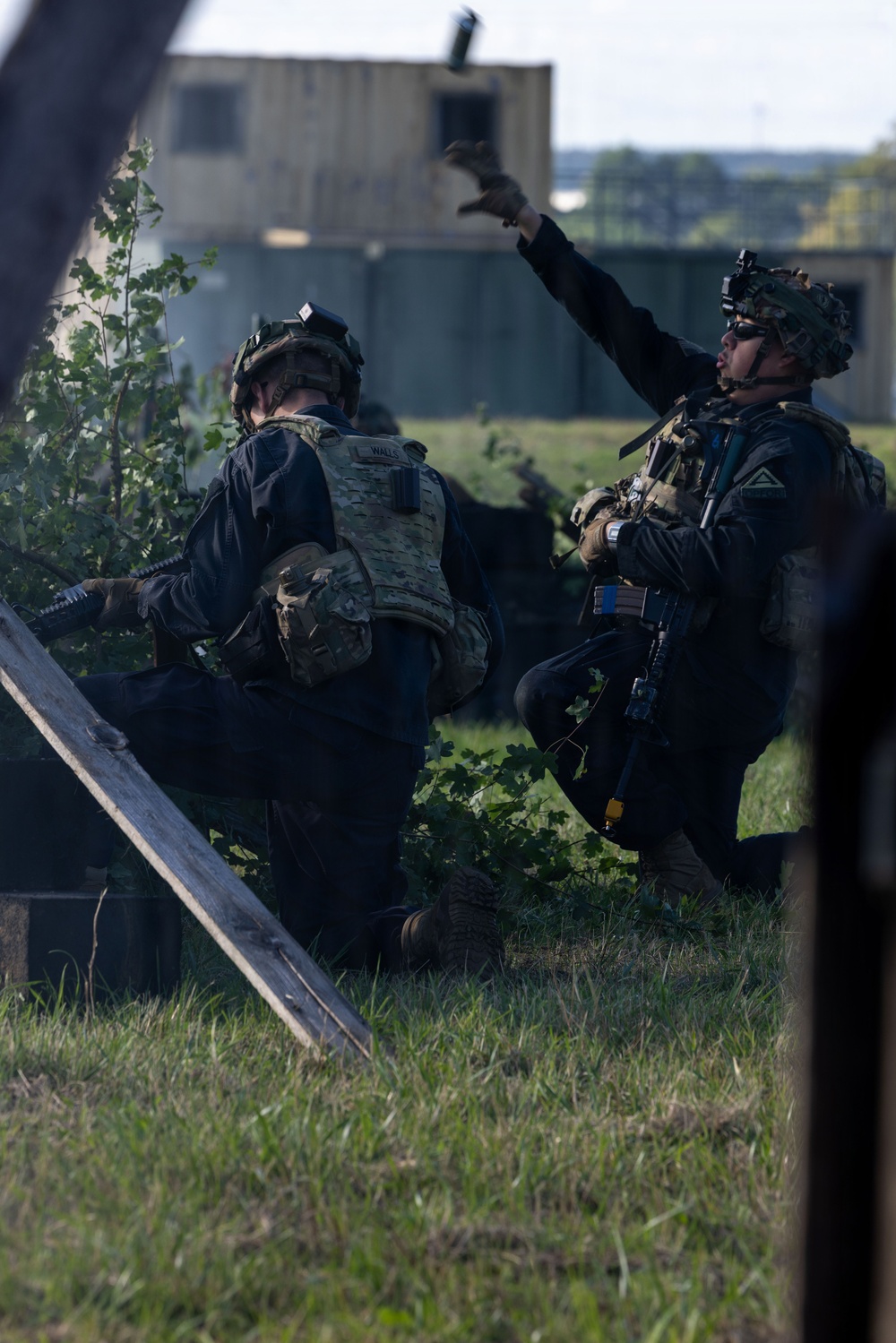 Two military personnel in tactical gear are positioned in a grassy area near what appears to be a training facility or urban operations environment. One soldier is kneeling while the other is in a standing/crouching position with arm raised, appearing to throw an object — likely a simulated grenade or training device. The setting and equipment suggest a military training exercise rather than a combat operation.