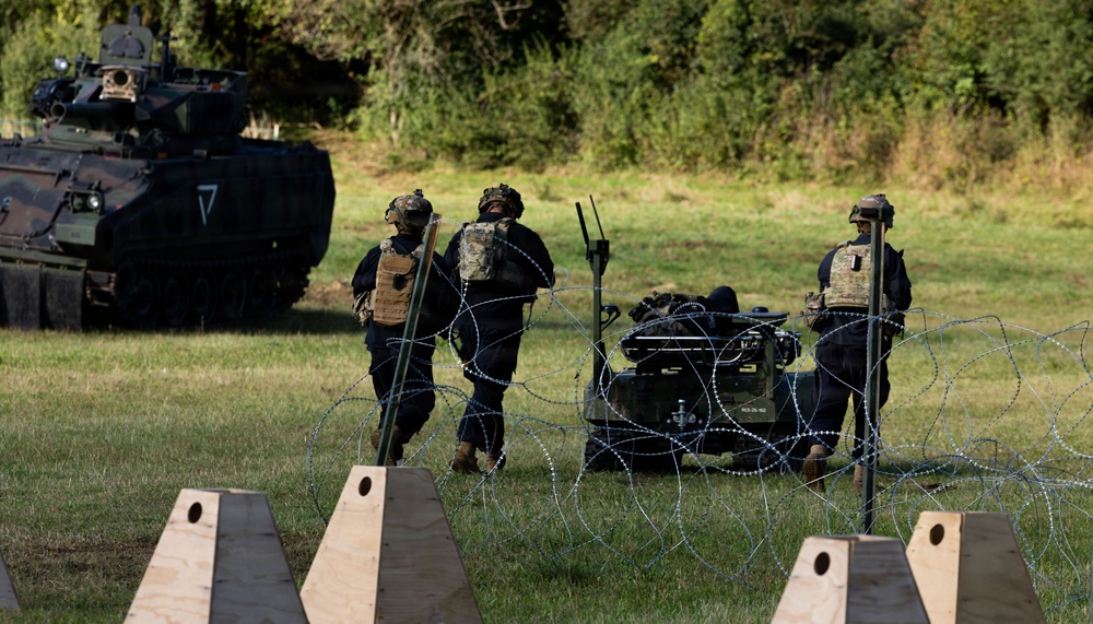 Military personnel in black uniforms with tactical gear are working near concertina wire obstacles and a ground robot or unmanned vehicle on a grassy field. A Bradley Fighting Vehicle (or similar armored personnel carrier) is visible in the background on the left. The scene appears to depict a military training exercise involving obstacle breaching and unmanned ground systems.