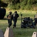 Military personnel in black uniforms with tactical gear are working near concertina wire obstacles and a ground robot or unmanned vehicle on a grassy field. A Bradley Fighting Vehicle (or similar armored personnel carrier) is visible in the background on the left. The scene appears to depict a military training exercise involving obstacle breaching and unmanned ground systems.