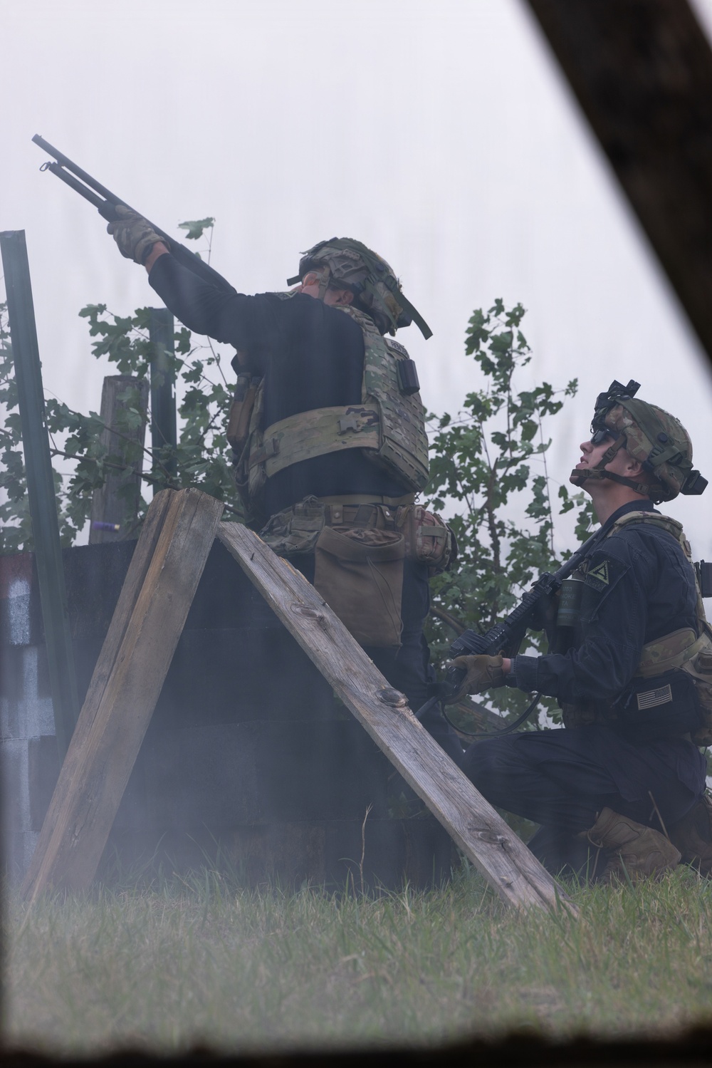Two military personnel in tactical gear are positioned near a wooden structure amid smoke or haze. The standing individual is aiming a shotgun upward while the kneeling individual holds an assault rifle and observes upward. Both wear multicam/OCP pattern helmets, body armor, and dark uniforms consistent with U.S. Army equipment.