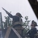 Two military personnel in tactical gear are positioned near a wooden structure amid smoke or haze. The standing individual is aiming a shotgun upward while the kneeling individual holds an assault rifle and observes upward. Both wear multicam/OCP pattern helmets, body armor, and dark uniforms consistent with U.S. Army equipment.