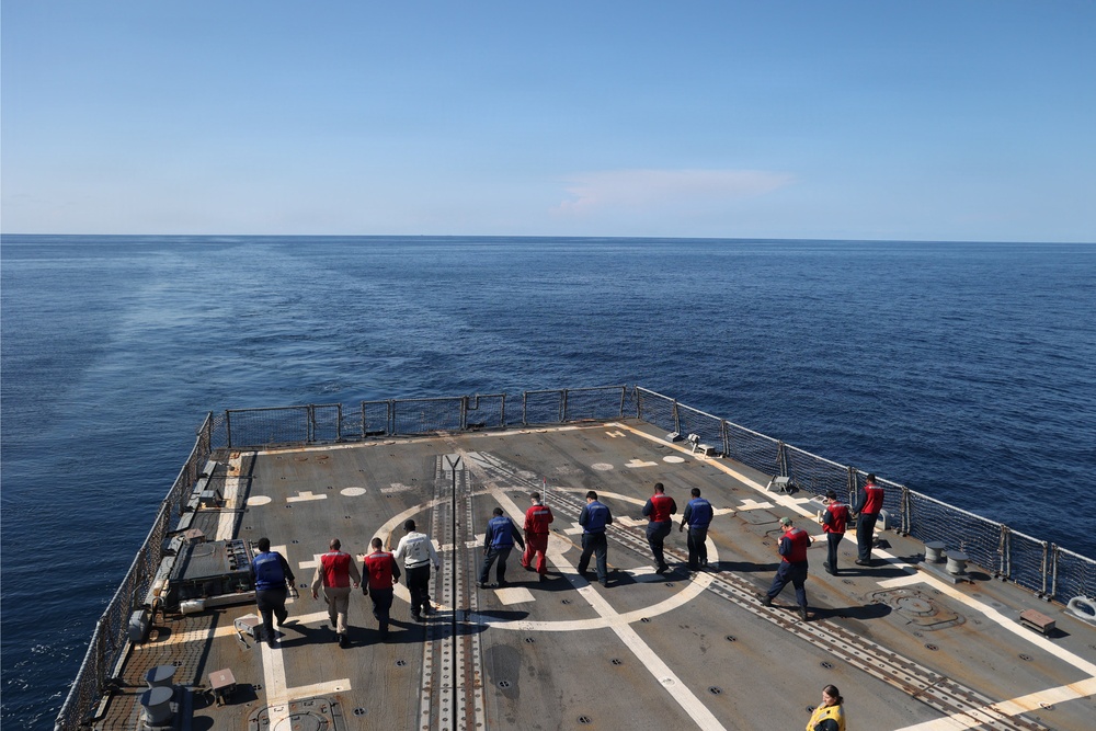 The image shows a group of approximately 12-15 personnel walking on what appears to be a helicopter landing deck (helideck) of a naval or coast guard vessel at sea. The deck features standard helipad markings including an 'H' symbol and circular landing guides. The vessel is underway on calm, open blue ocean under clear skies.