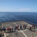 The image shows a group of approximately 12-15 personnel walking on what appears to be a helicopter landing deck (helideck) of a naval or coast guard vessel at sea. The deck features standard helipad markings including an 'H' symbol and circular landing guides. The vessel is underway on calm, open blue ocean under clear skies.