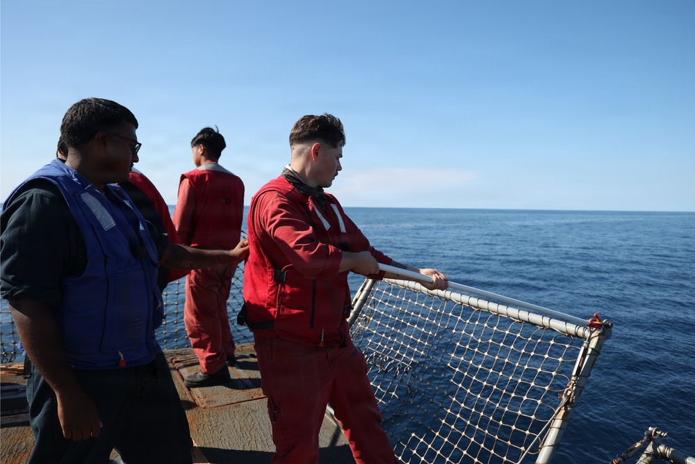 Three individuals are working on the deck of a vessel at sea, wearing red coverall suits and personal flotation devices. The foreground figure is handling a metal-framed safety net or gate along the ship's railing. The scene takes place under clear blue skies with calm open ocean visible in the background.