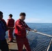 Three individuals are working on the deck of a vessel at sea, wearing red coverall suits and personal flotation devices. The foreground figure is handling a metal-framed safety net or gate along the ship's railing. The scene takes place under clear blue skies with calm open ocean visible in the background.