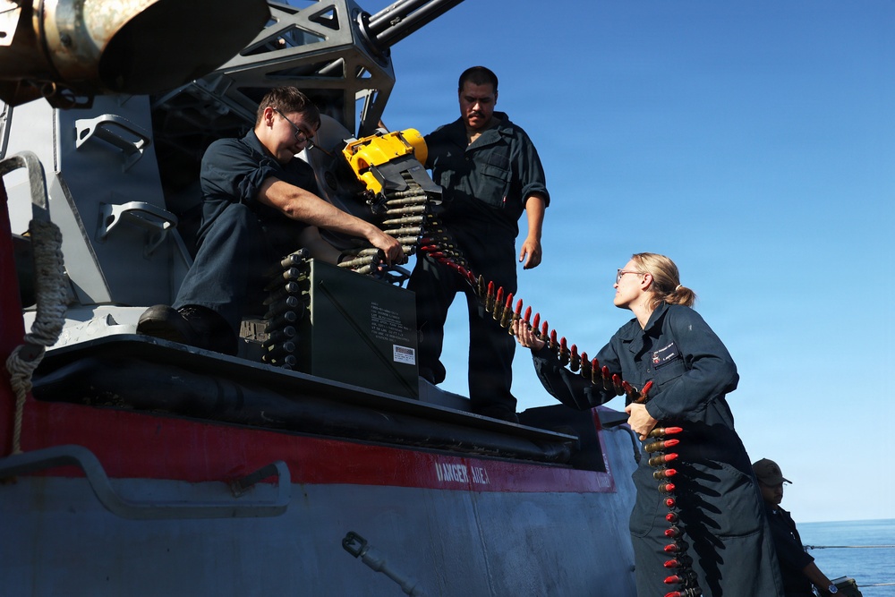 Three naval personnel in dark blue uniforms are loading an ammunition belt into a deck-mounted weapon system aboard a military vessel at sea. One sailor is handling the ammunition chain from the deck level while another feeds it into the weapon mount from an elevated position, with a third crew member observing. A 'DANGER AREA' warning is visibly stenciled on the gun mount housing.