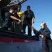 Three naval personnel in dark blue uniforms are loading an ammunition belt into a deck-mounted weapon system aboard a military vessel at sea. One sailor is handling the ammunition chain from the deck level while another feeds it into the weapon mount from an elevated position, with a third crew member observing. A 'DANGER AREA' warning is visibly stenciled on the gun mount housing.