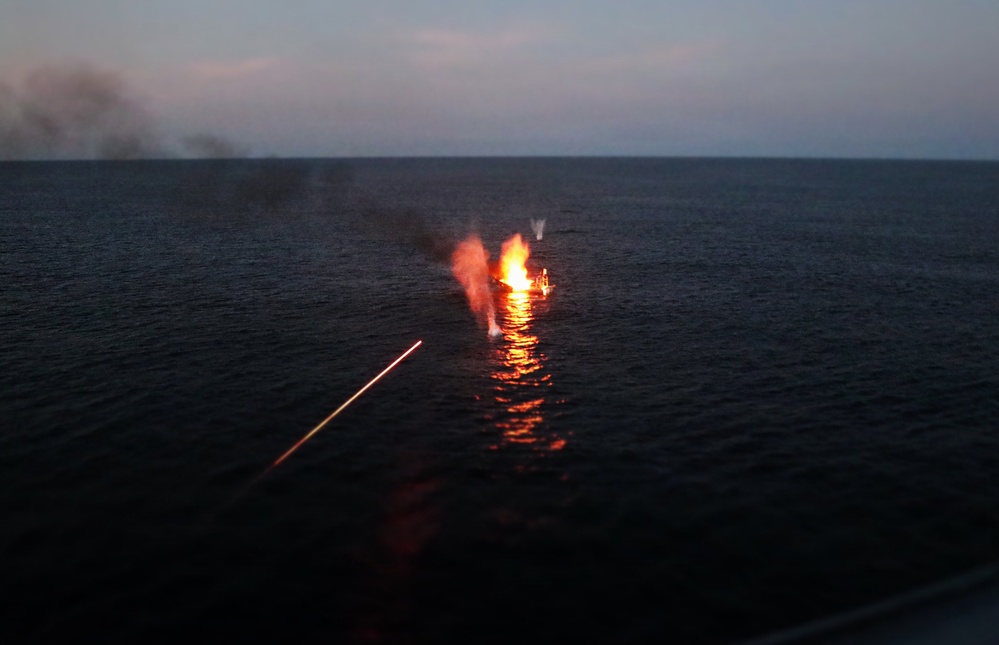 A vessel or object on open water is engulfed in flames at dusk or dawn, with visible fire, smoke plumes, and water splashes surrounding it. A bright diagonal streak of light — consistent with a tracer round or laser designator — is visible in the foreground, suggesting an active engagement or weapons fire. Dark smoke drifts toward the upper left horizon, indicating the fire has been burning for some time.
