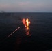 A vessel or object on open water is engulfed in flames at dusk or dawn, with visible fire, smoke plumes, and water splashes surrounding it. A bright diagonal streak of light — consistent with a tracer round or laser designator — is visible in the foreground, suggesting an active engagement or weapons fire. Dark smoke drifts toward the upper left horizon, indicating the fire has been burning for some time.