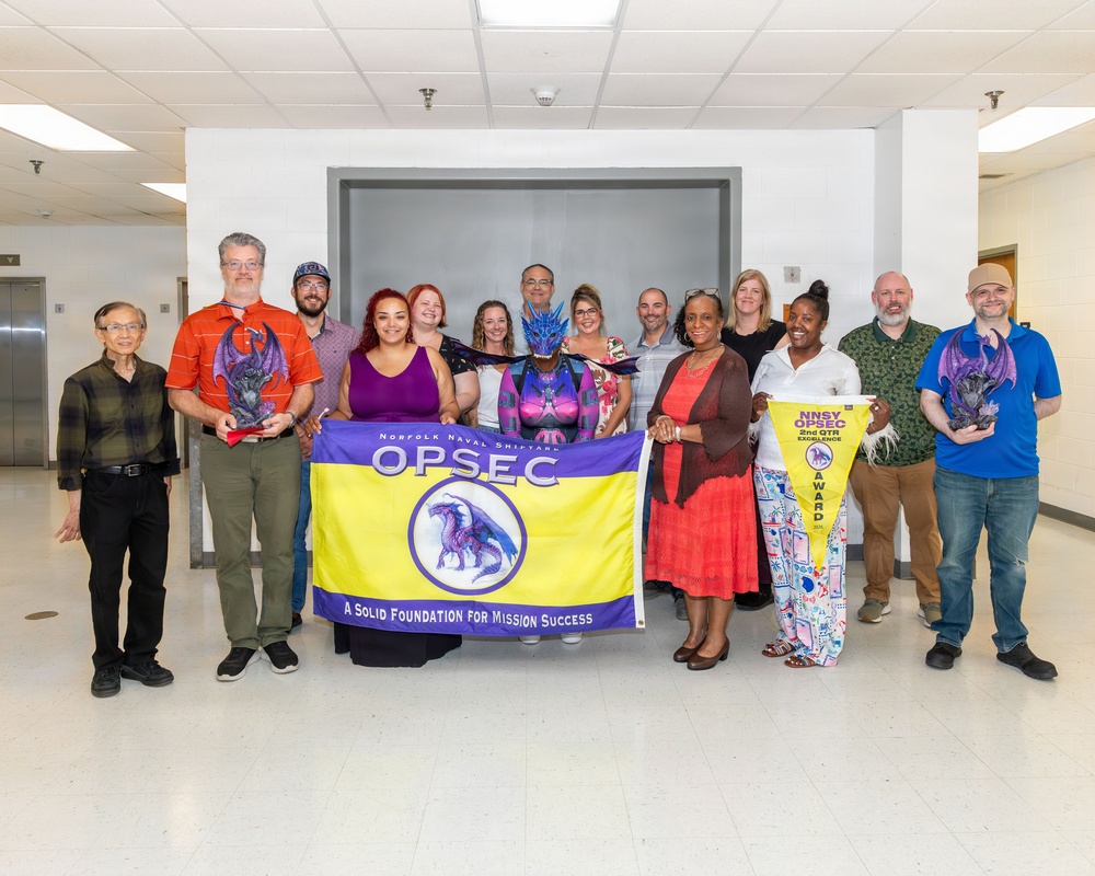 A group of approximately 14 people pose for a formal group photo in what appears to be a government or military facility hallway. They are gathered around a yellow and purple banner reading 'Norfolk Naval Shipyard OPSEC - A Solid Foundation for Mission Success.' Several individuals are holding dragon figurines and a pennant award.
