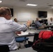 A group of adults are gathered in a modern classroom or conference room setting, appearing to be in a meeting or training session. A man in a light blue dress shirt is seated in the foreground with his back to the camera, gesturing toward others in the room. Several other participants are seated at tables facing the front of the room.