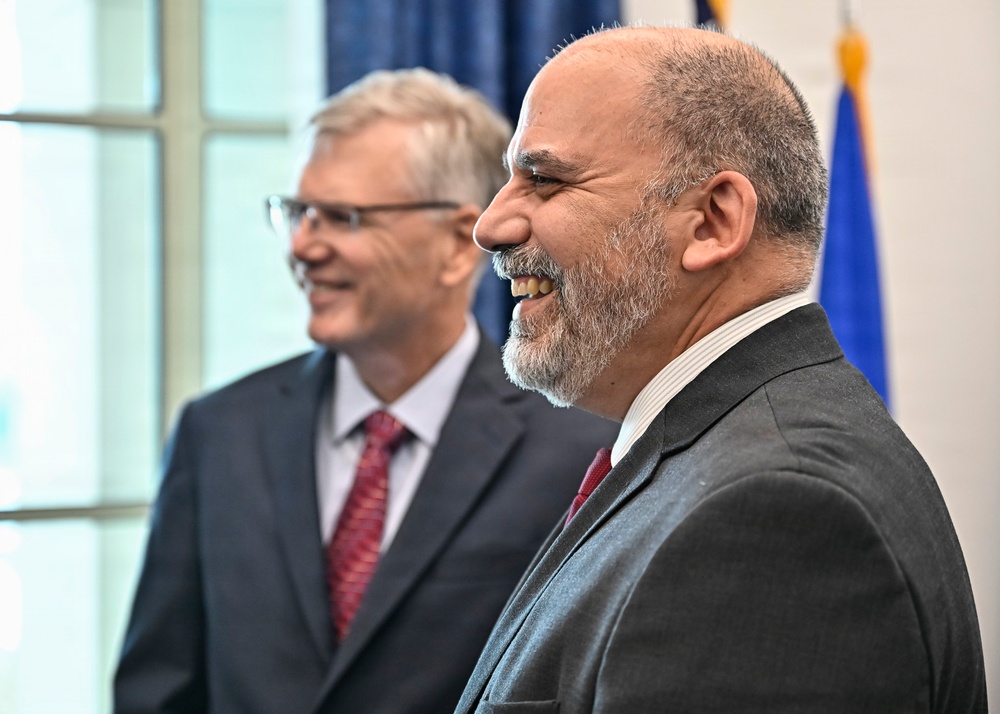 Two men in formal business attire are standing together in what appears to be an official or governmental setting, both smiling. The foreground subject is a bald man with a gray beard wearing a dark gray suit and burgundy tie, while the background subject is an older man with gray hair and glasses wearing a dark suit with a red patterned tie. A flag, likely American, is partially visible in the background.