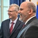 Two men in formal business attire are standing together in what appears to be an official or governmental setting, both smiling. The foreground subject is a bald man with a gray beard wearing a dark gray suit and burgundy tie, while the background subject is an older man with gray hair and glasses wearing a dark suit with a red patterned tie. A flag, likely American, is partially visible in the background.